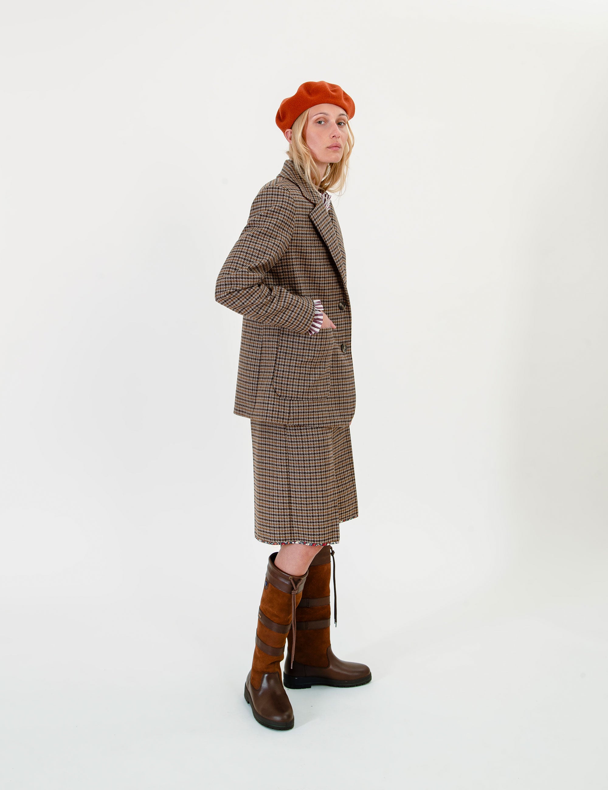 A woman stands in profile wearing a brown and beige plaid blazer and matching skirt set, an orange-red beret, and tall brown leather boots against a plain white studio background.