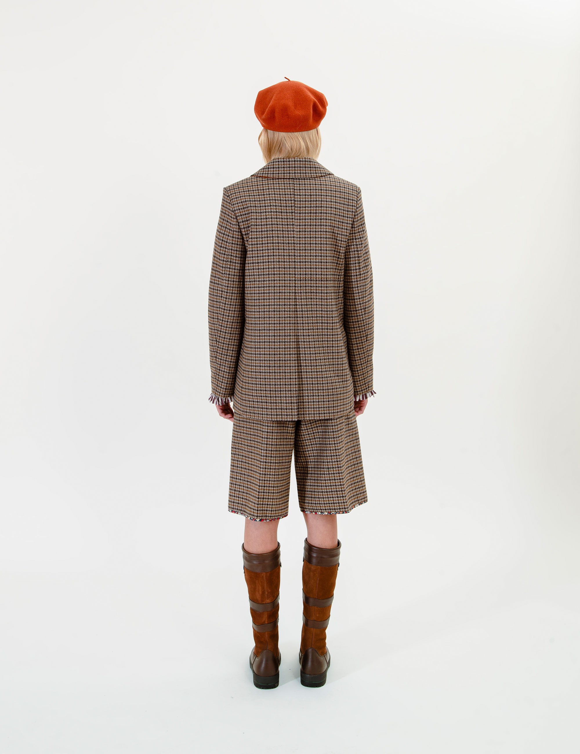 A woman shown from the back wearing a brown and beige checkered blazer and matching shorts, paired with an orange beret and brown knee-high boots, against a plain white studio background.