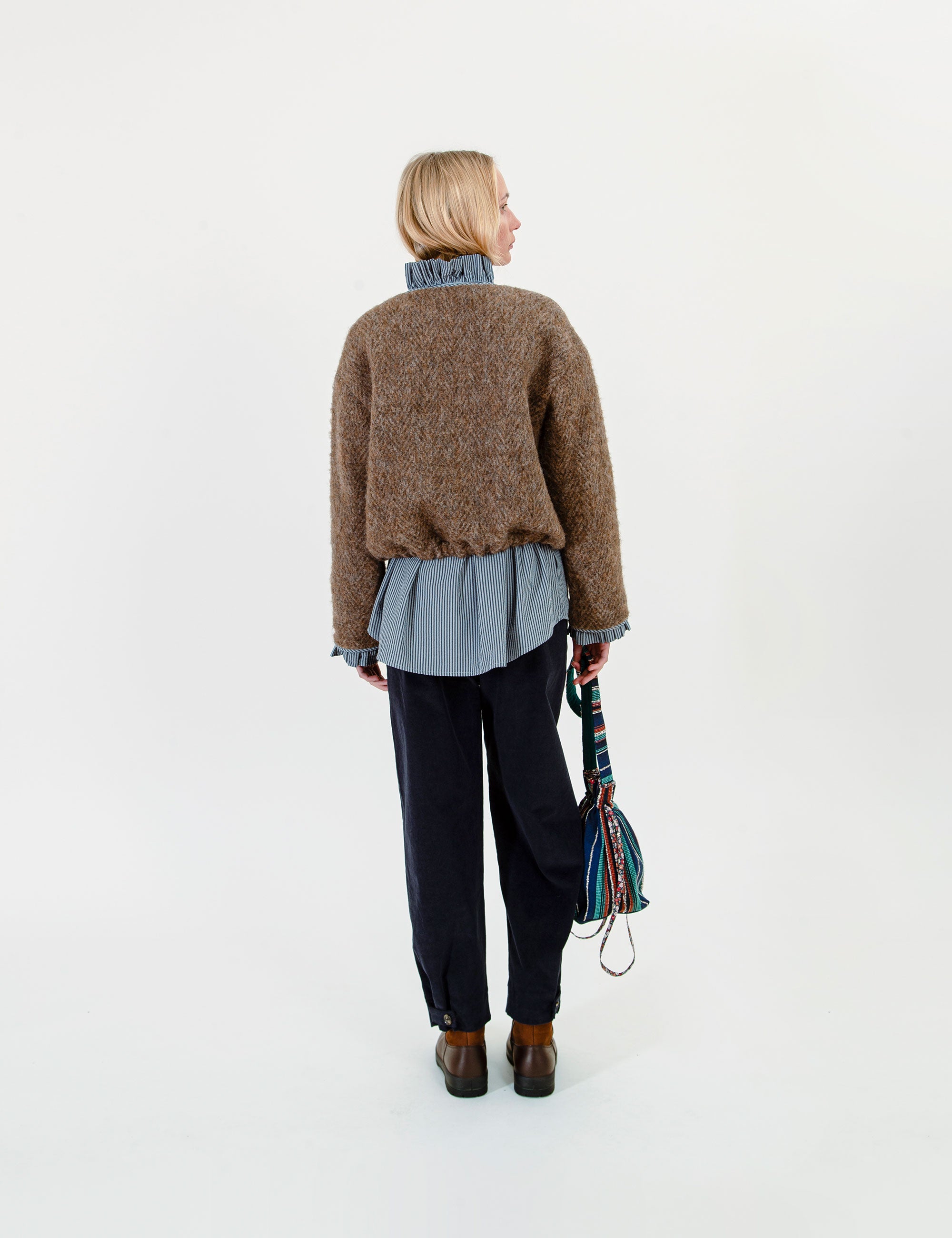 Woman wearing a brown sweater over a blue shirt with dark pants, holding a colorful bag on a white background