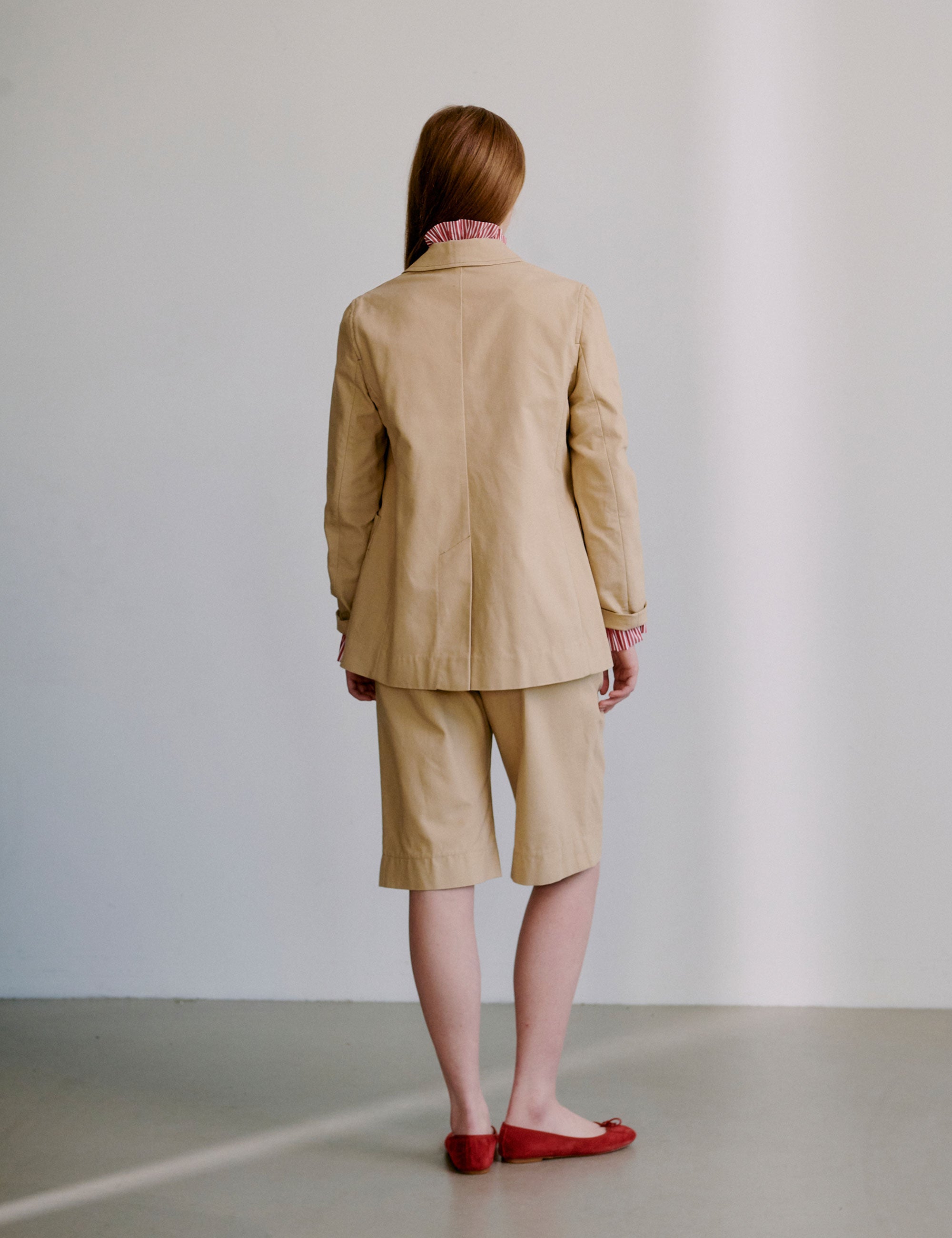 Woman wearing a beige jacket with a red patterned shirt underneath and matching shorts, standing against a plain background.