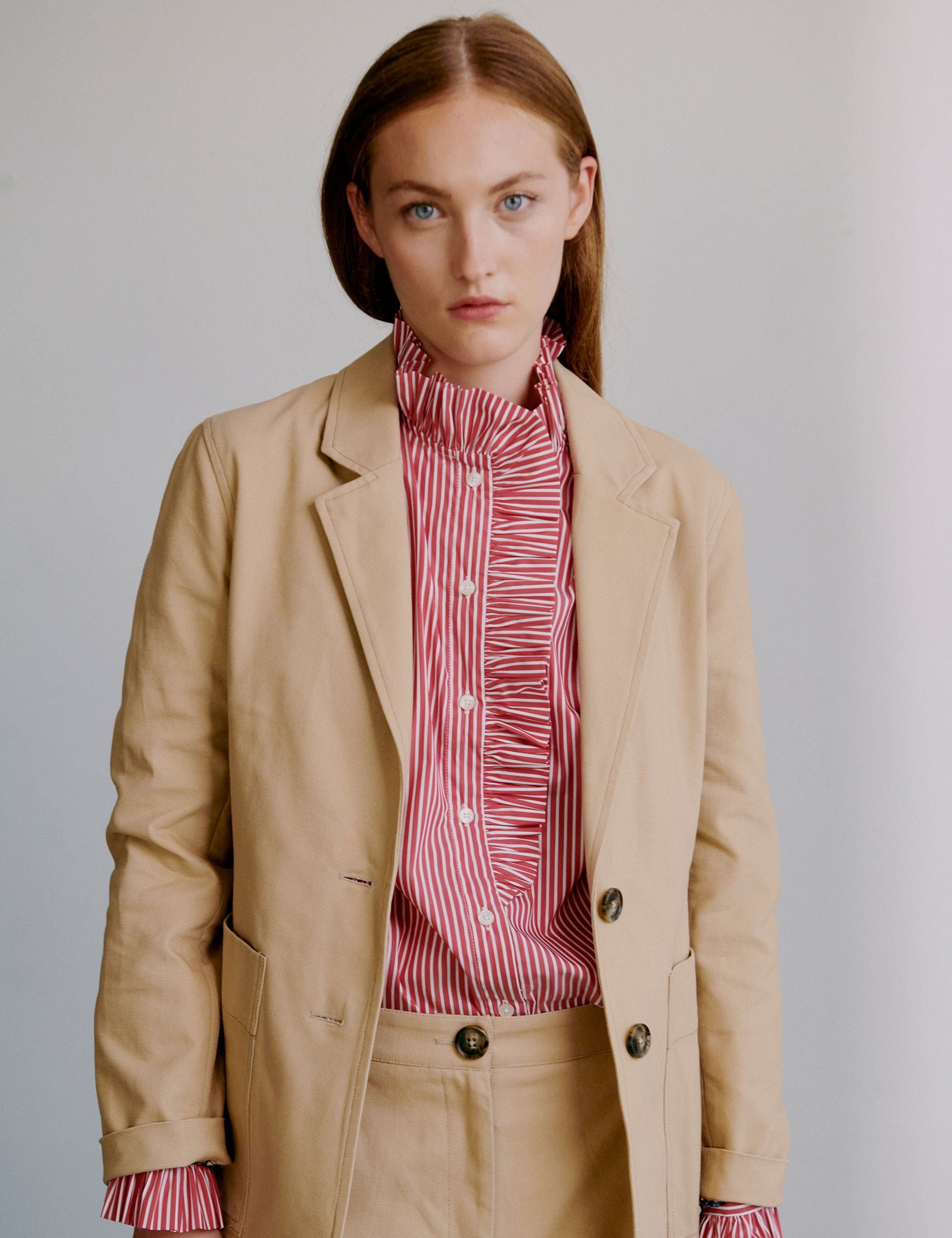 Woman wearing a beige jacket with a red patterned shirt underneath and matching shorts, standing against a plain background.