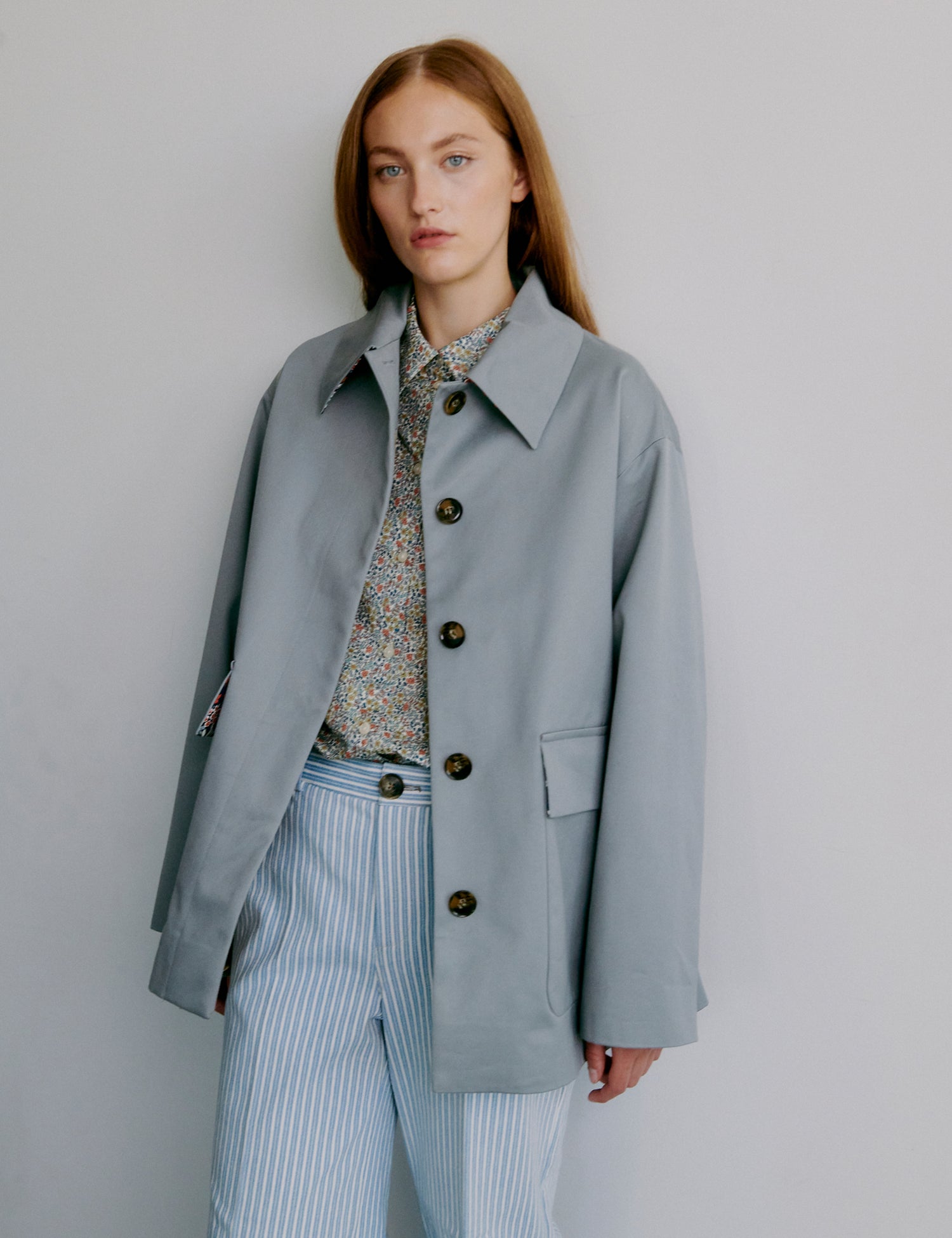 Woman wearing a light gray coat over a patterned shirt and striped pants against a plain background