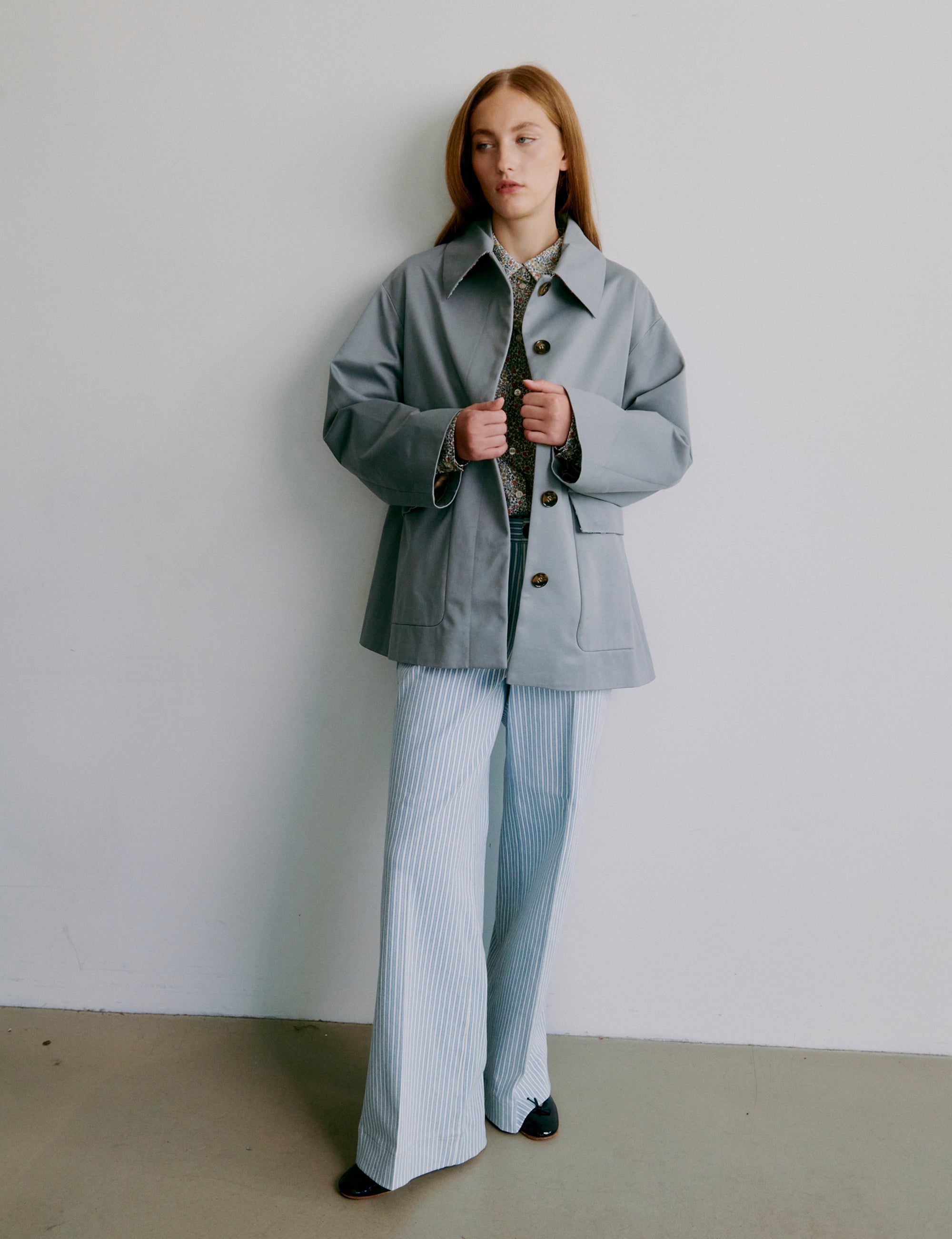 Kvinna i blommig långärmad skjorta och ljusblårandiga byxor mot neutral bakgrund