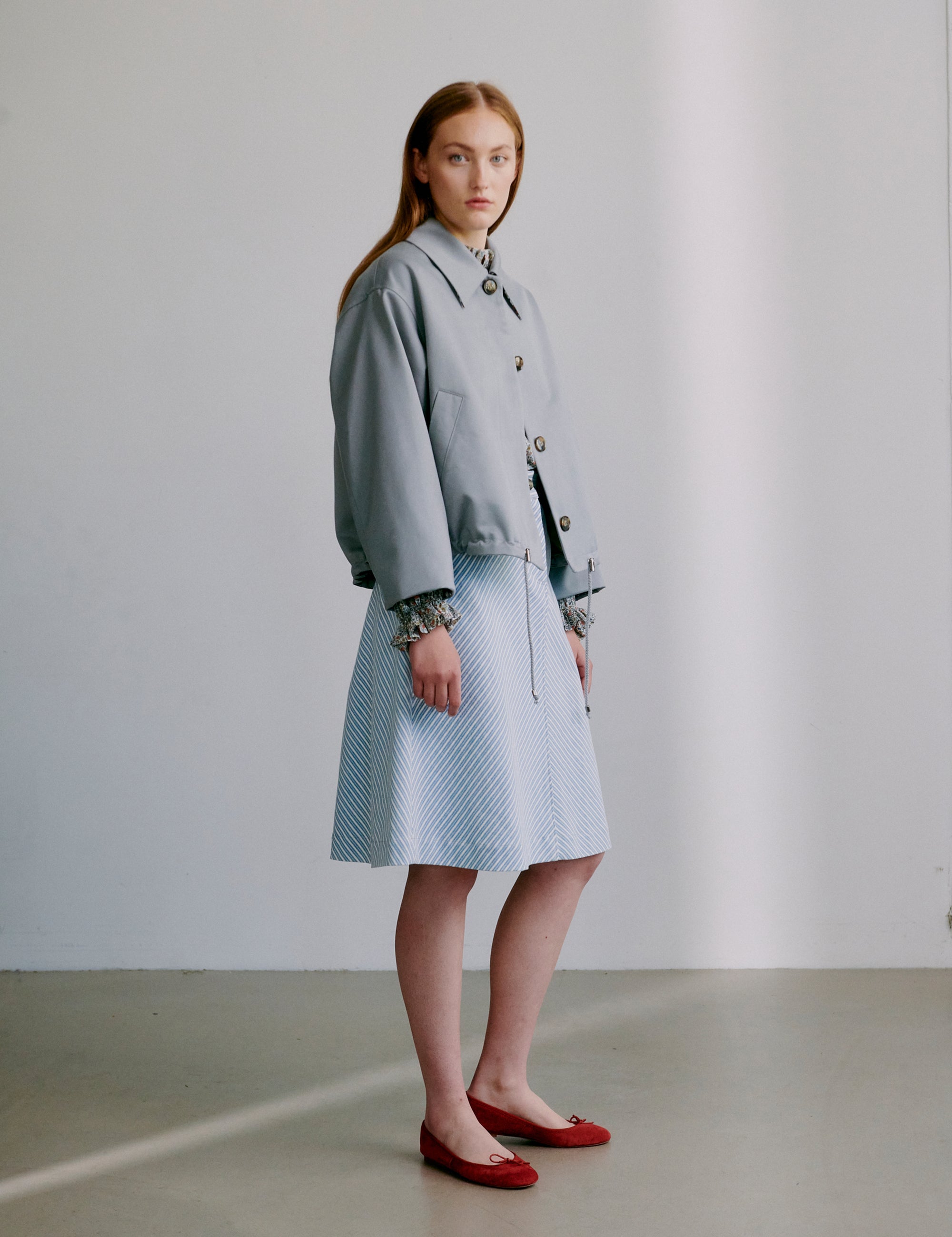 Woman wearing a light grey jacket and blue and white striped skirt with red shoes against a plain background