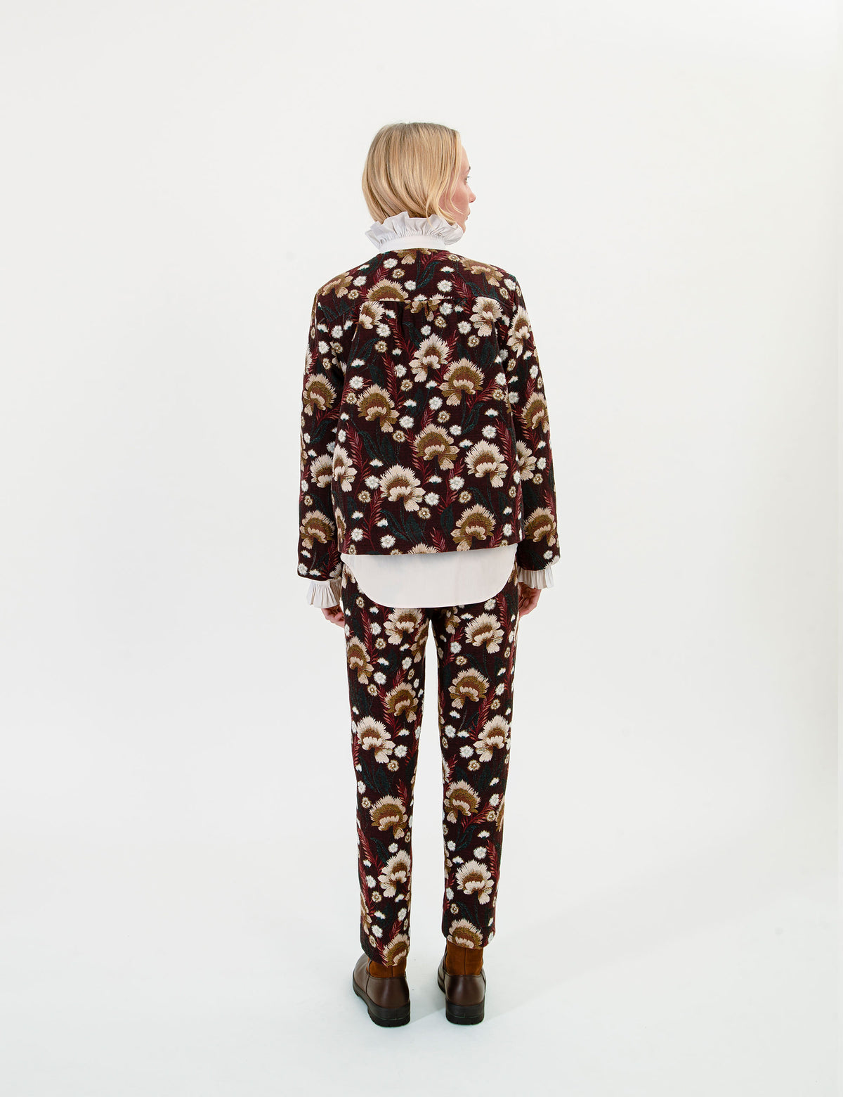 A woman shown from the back wearing a matching two-piece suit with a dark brown base and a beige and white floral pattern, styled with a white ruffled shirt and brown leather shoes against a plain white studio background.