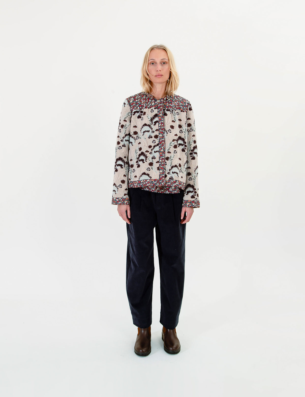 A woman wearing a cream patterned jacket with dark trim, dark trousers, and brown boots against a plain white studio background.