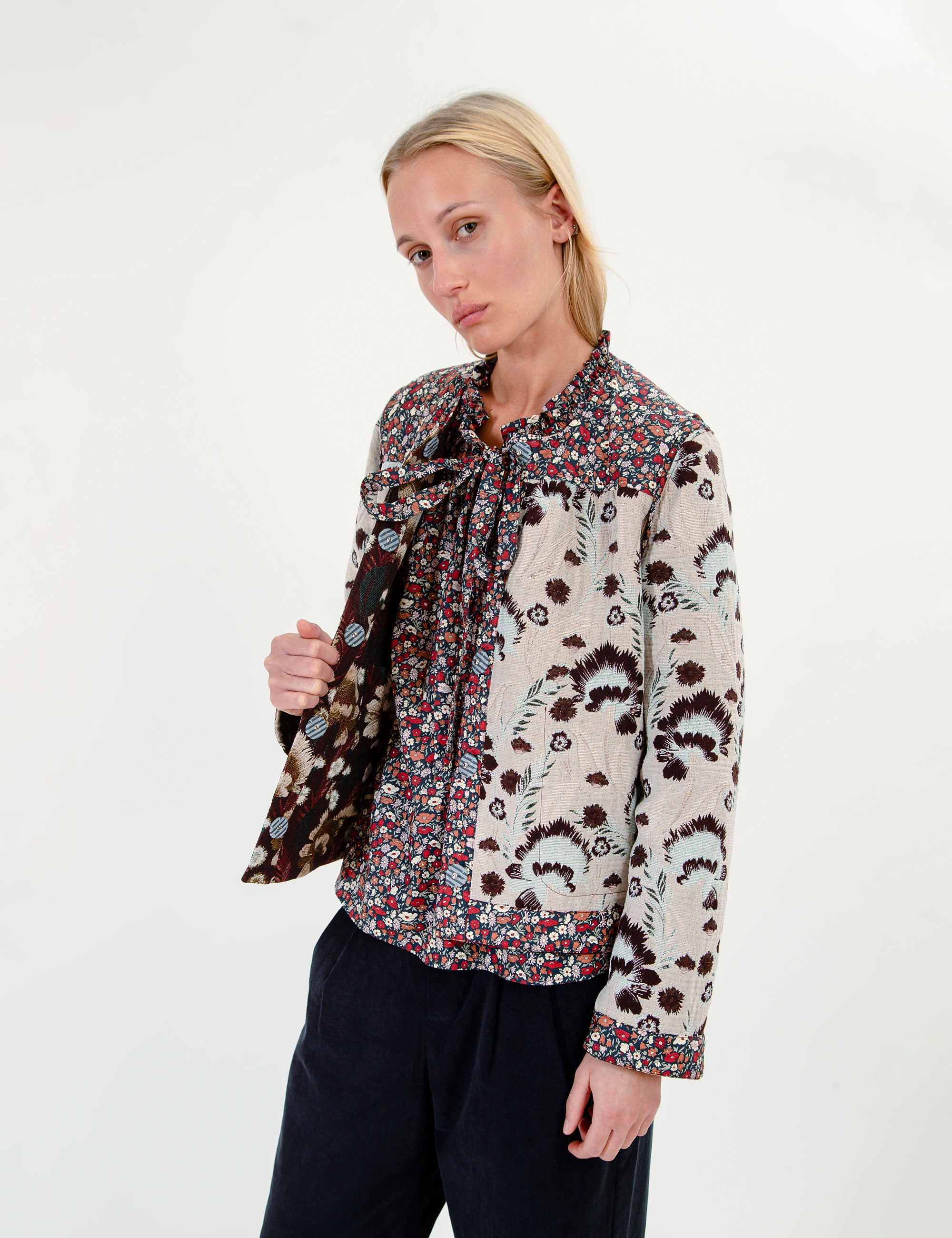 A woman modeling a long-sleeve blouse with a tie-neck and a dense abstract floral pattern in cream, brown, red, and blue, paired with dark trousers against a plain white studio background.