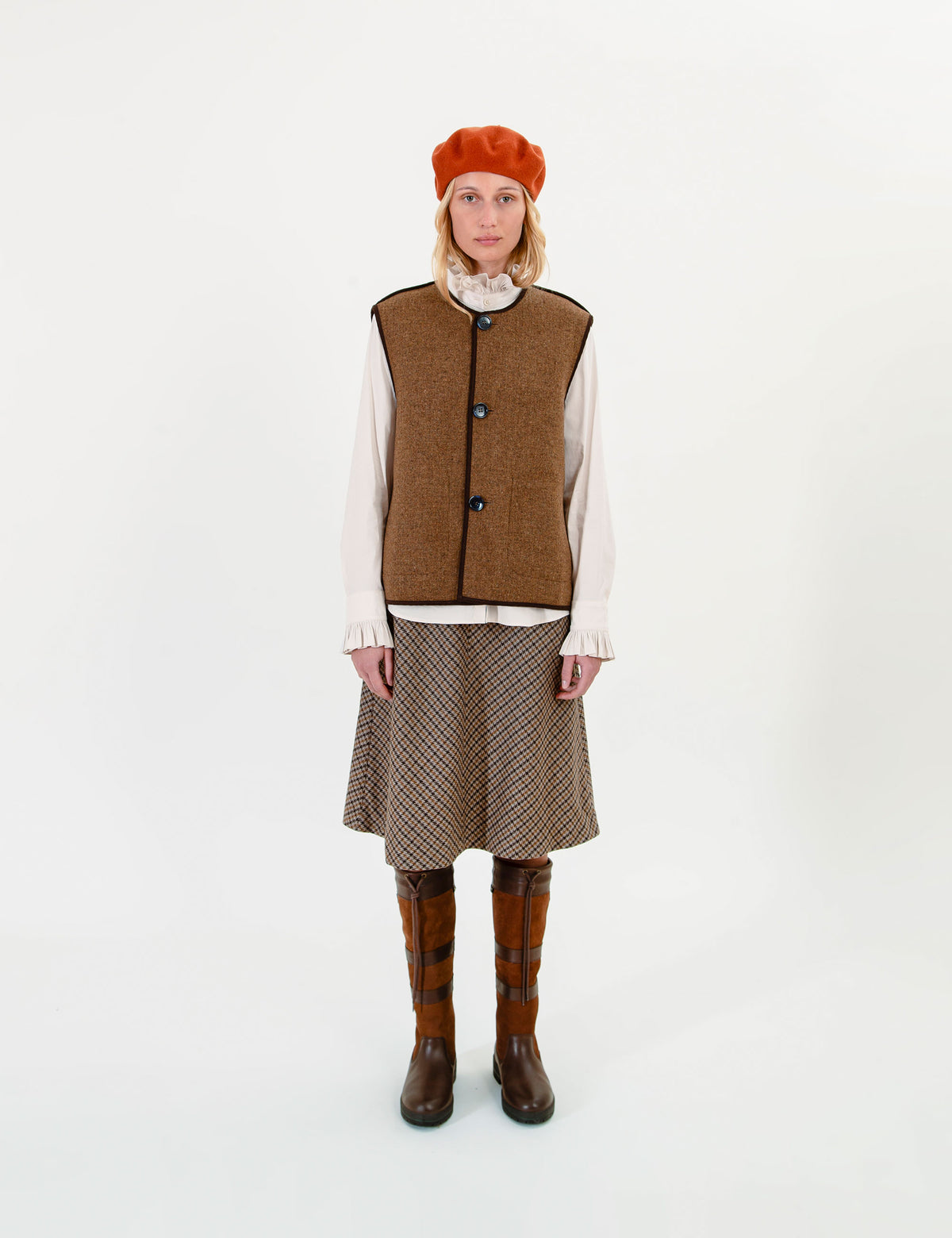 A woman stands against a plain white studio background wearing a brown tweed vest over a cream blouse, a brown patterned skirt, brown plaid knee-high socks, and an orange beret.