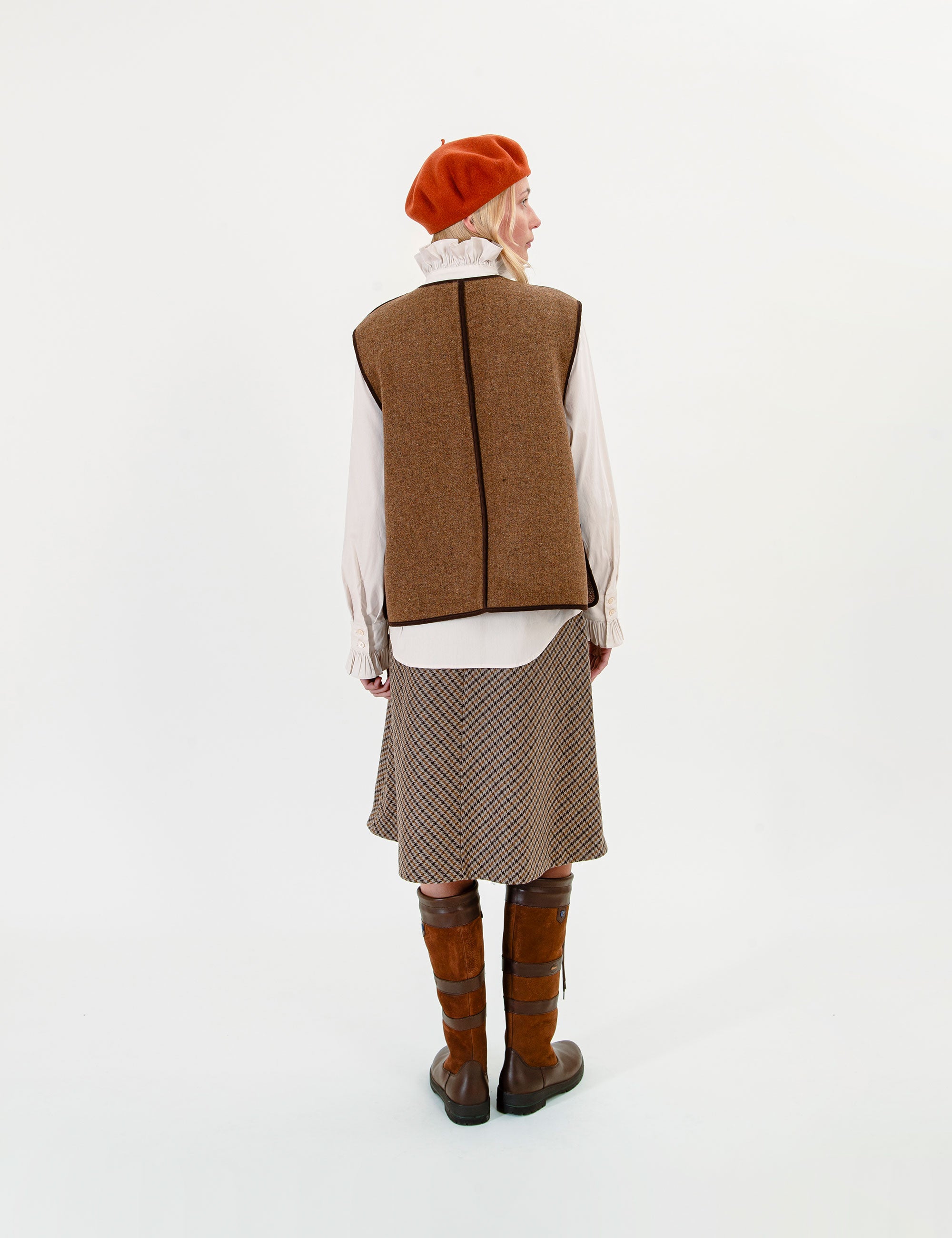 A woman shown from the back wearing a brown tweed vest over a white ruffled shirt, a brown patterned skirt, and brown leather boots, with a rust-orange beret on a white studio background.