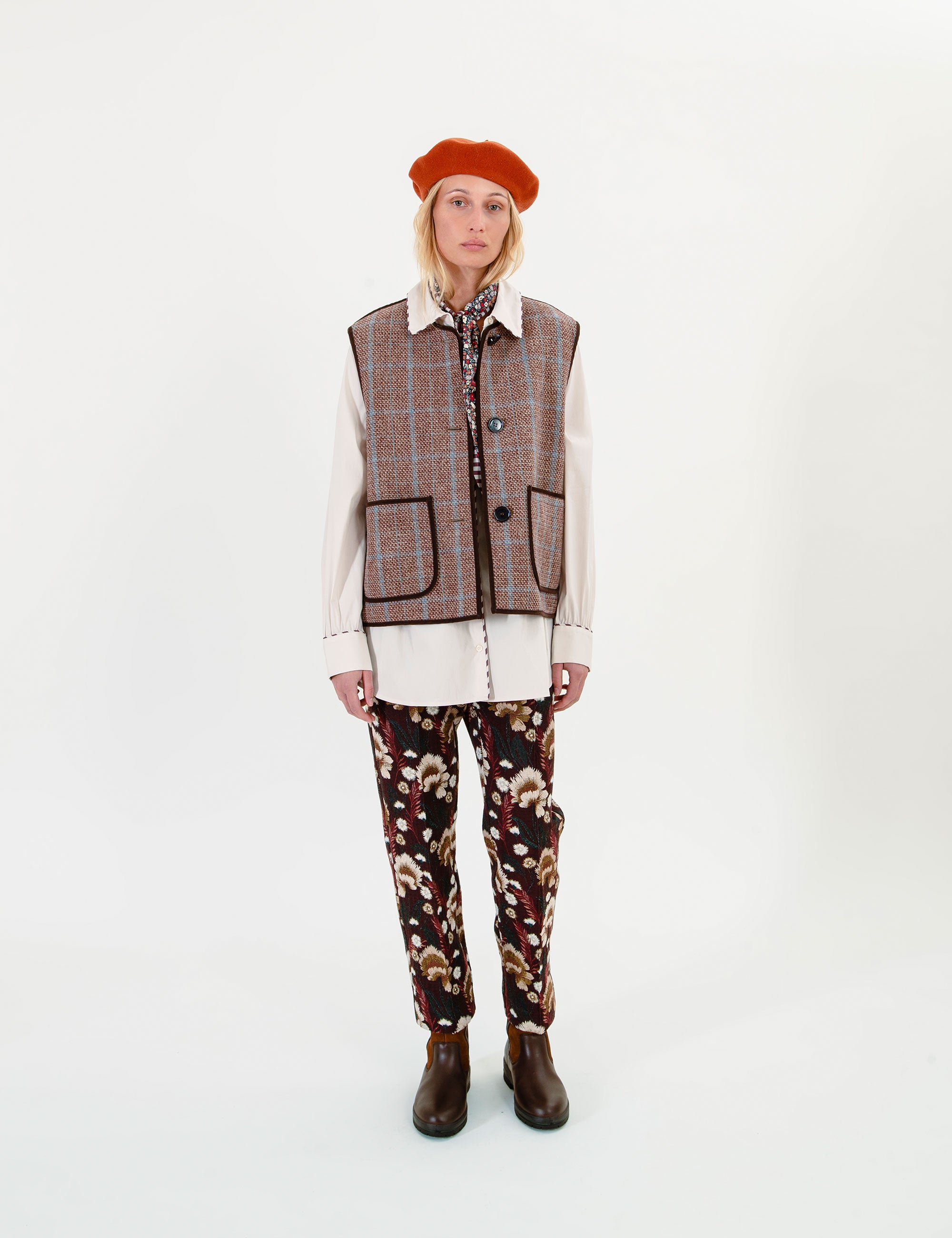 A woman stands against a plain white studio background wearing a brown plaid vest over a cream shirt, patterned pants, and a rust-orange beret.