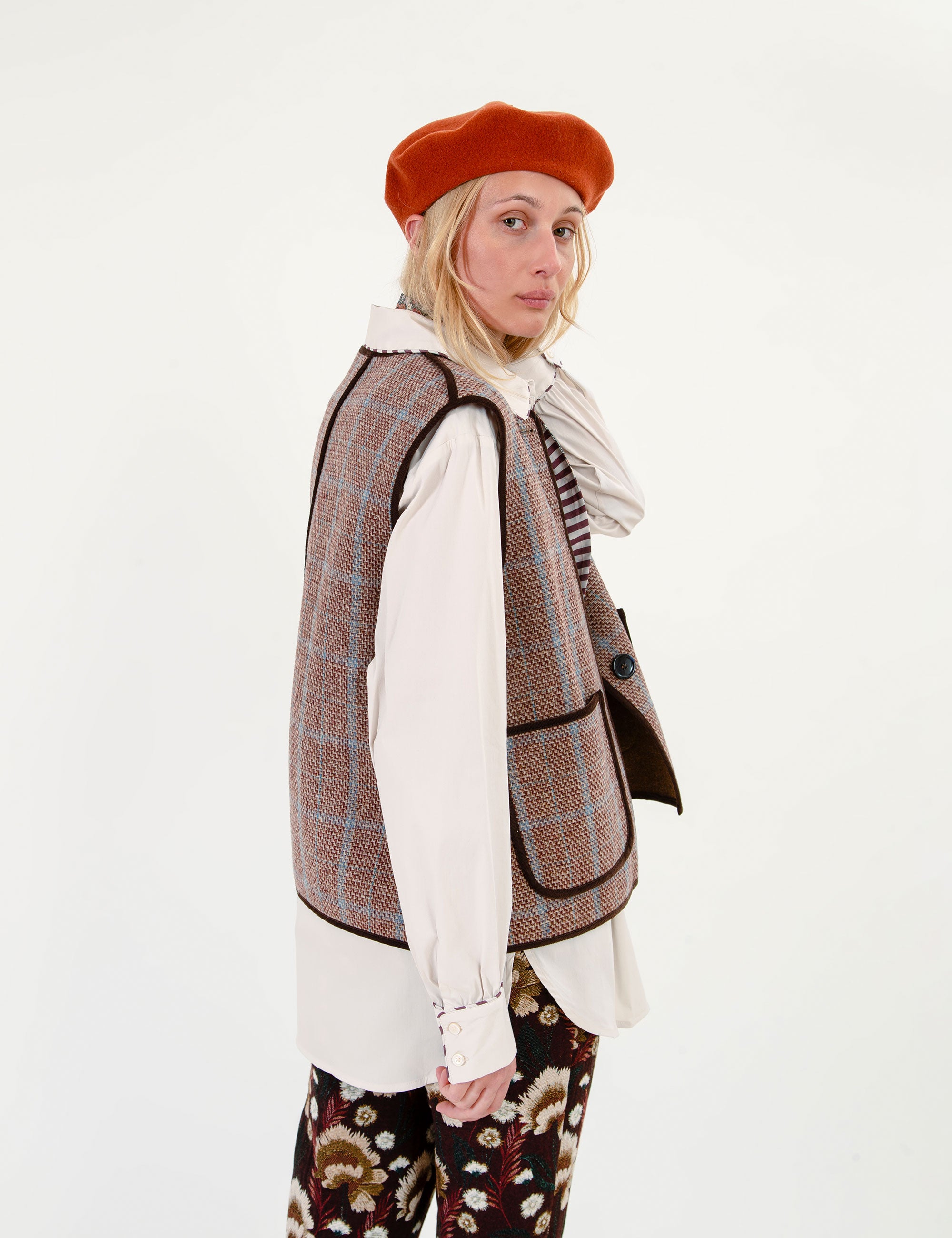 A woman wearing a brown and beige plaid tweed vest over a white ruffled blouse, paired with a rust-orange beret, a striped scarf, and patterned trousers, photographed against a plain white studio background.