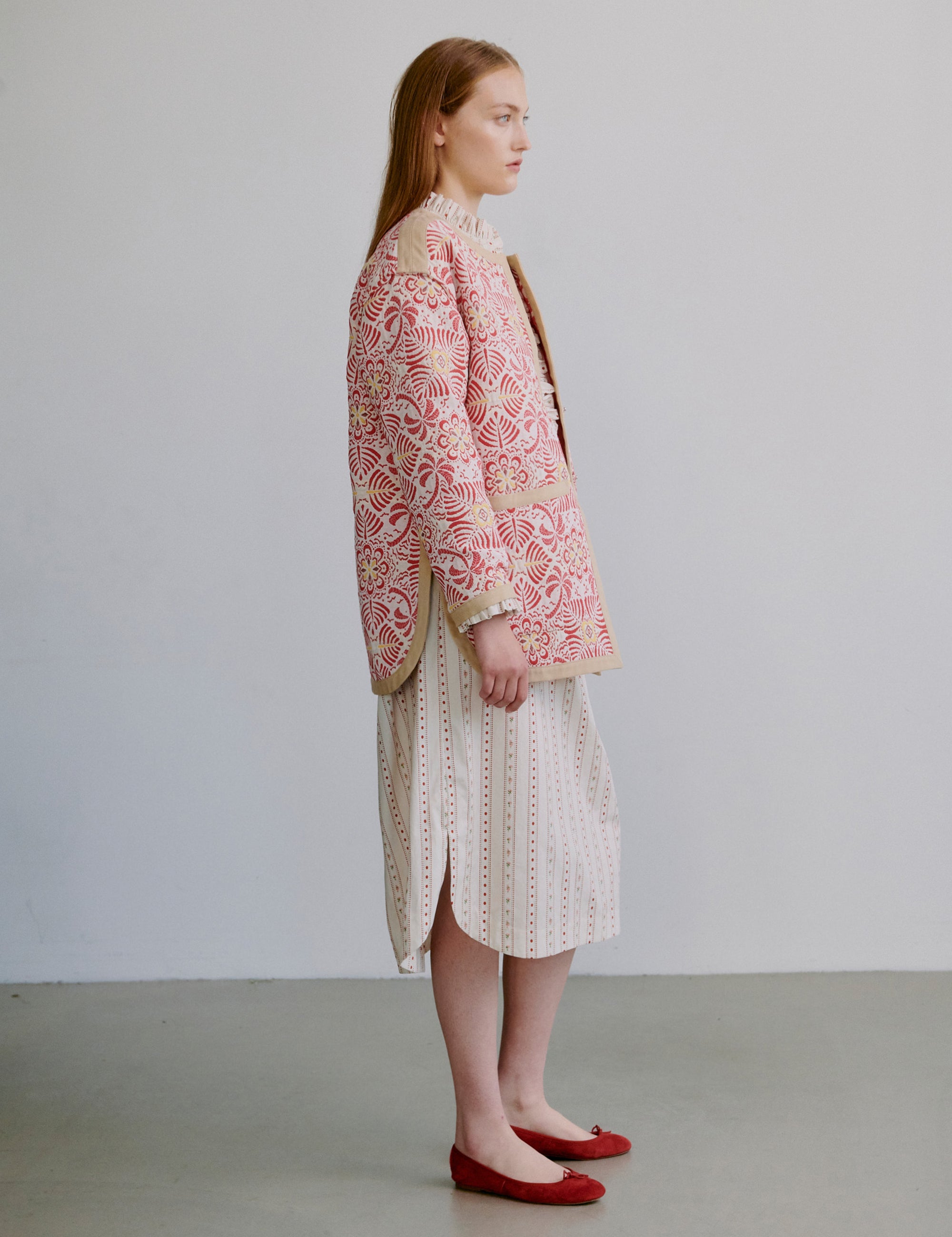 Woman wearing a red patterned jacket and creme white patterned dress against a plain background