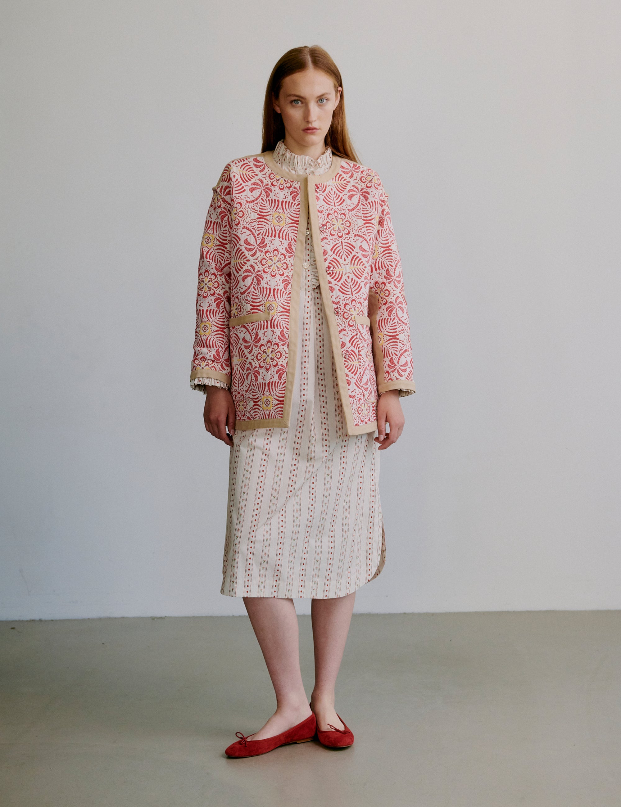 Woman wearing a red patterned jacket and creme white patterned dress against a plain background