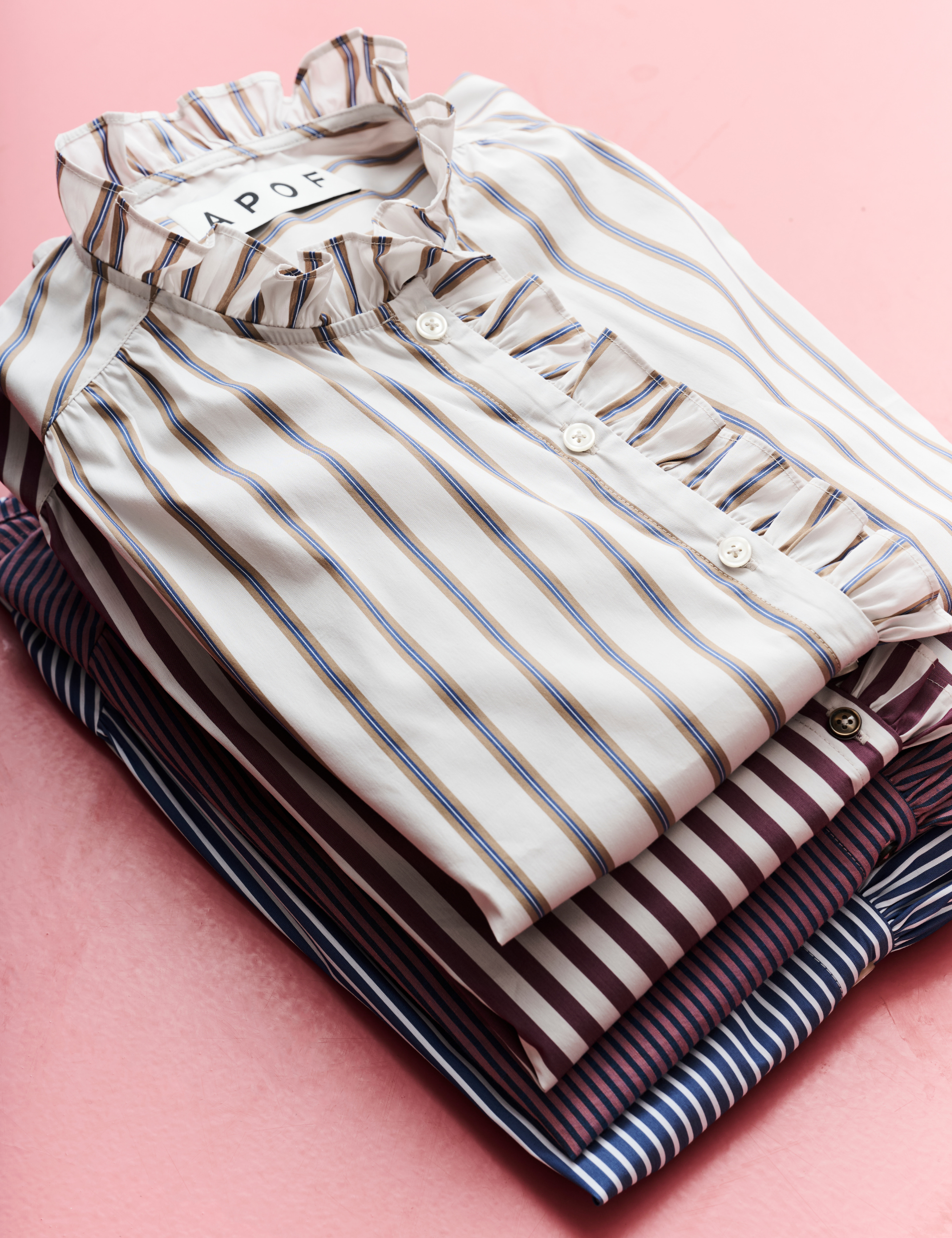 A stack of folded striped button-up shirts with ruffled trim, displayed on a pink background. The top shirt is cream with thin blue and brown stripes, and the shirts beneath show navy, burgundy, and dark blue striped patterns.