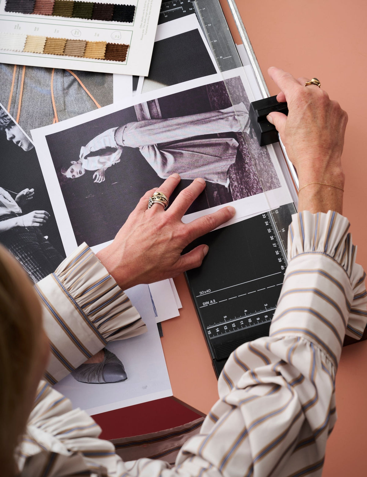 A close-up, over-the-shoulder view of a person's hands using a paper cutter with a black vinyl sheet on a beige striped long-sleeve blouse. The workspace is cluttered with fashion design elements, including color swatches, black-and-white fashion photography prints, and a ruler.