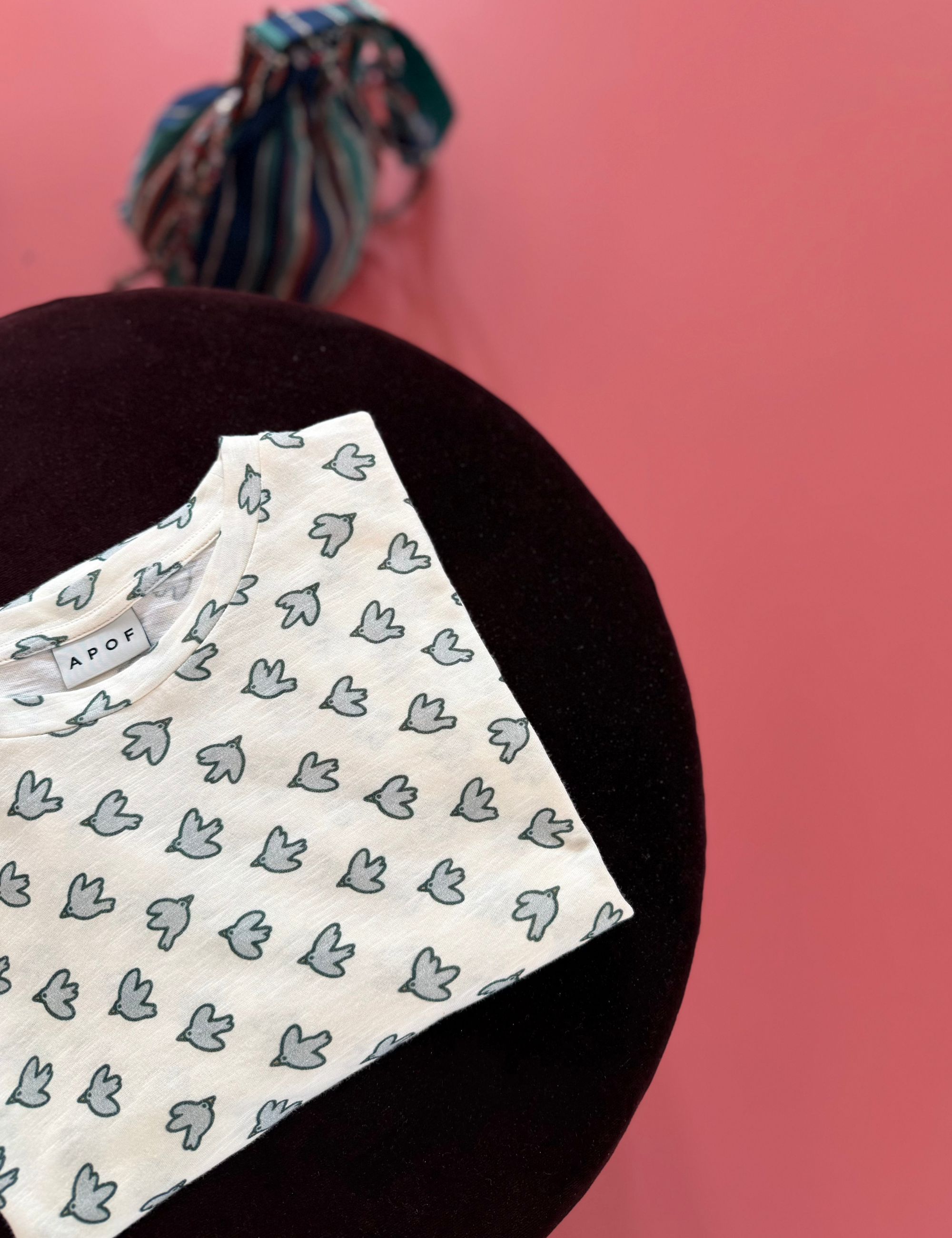 Printed long sleeve t-shirt with small bird motif, displayed on a dark velvet chair against a pink background.