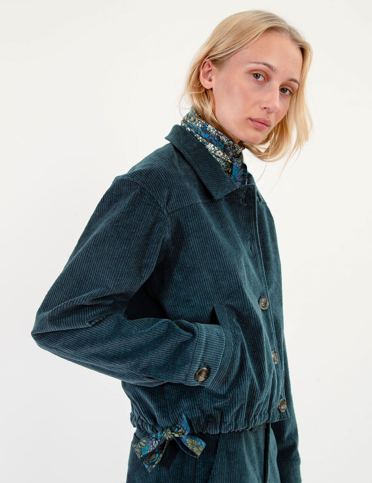 A woman wearing a dark teal corduroy jacket with a matching patterned neck scarf, photographed against a clean white studio background.
