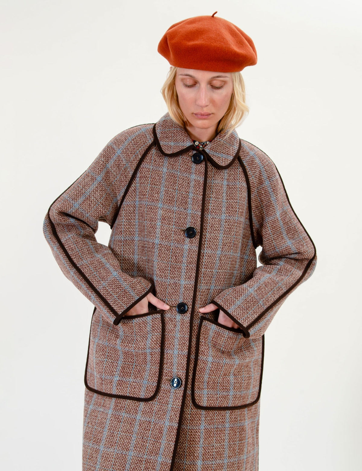 A woman wearing a brown and beige plaid coat with dark brown piping, paired with an orange beret, photographed against a plain off-white studio background.