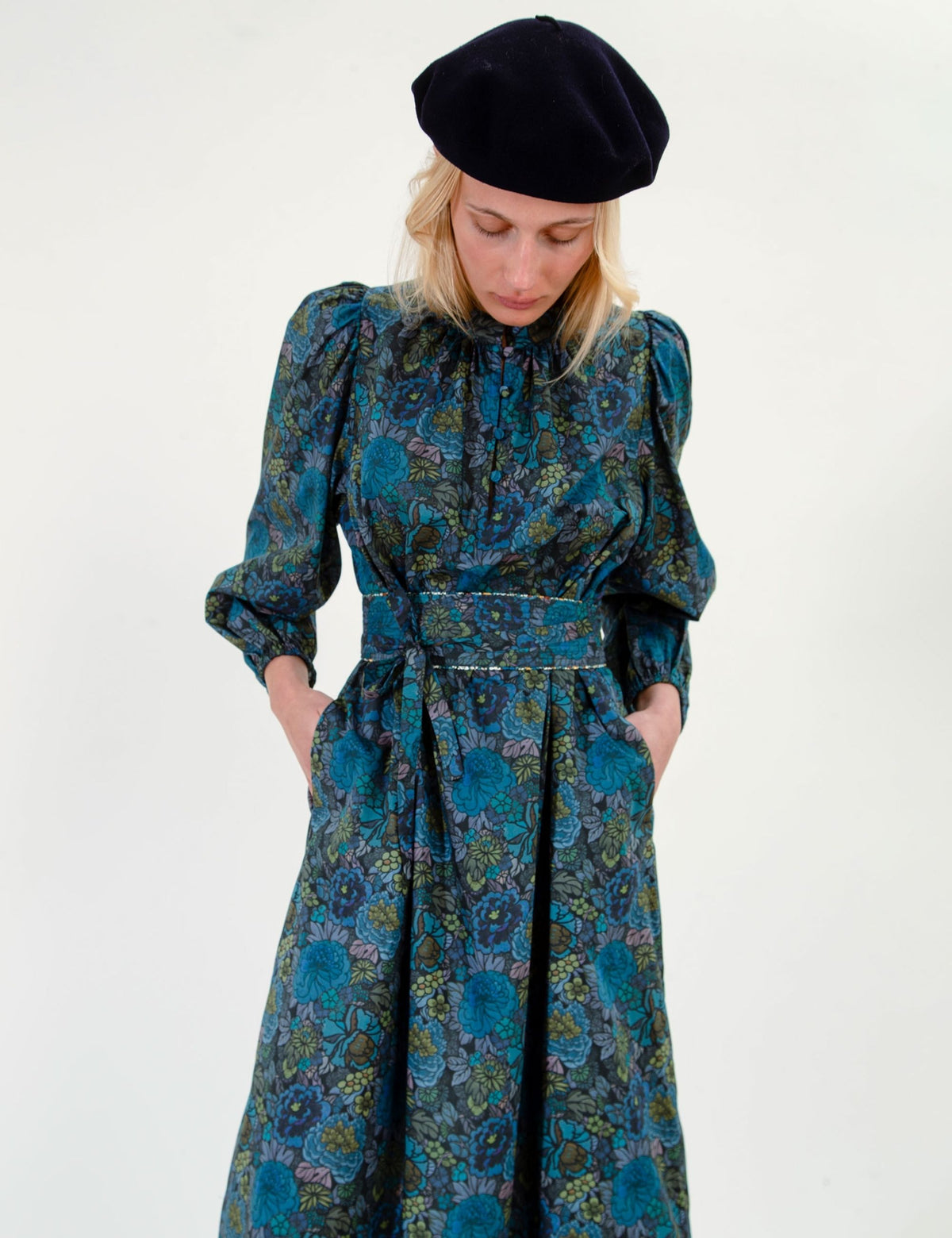 A woman wearing a long-sleeved, high-neck midi dress with a teal and blue floral pattern, paired with a black beret, photographed against a plain white studio background.