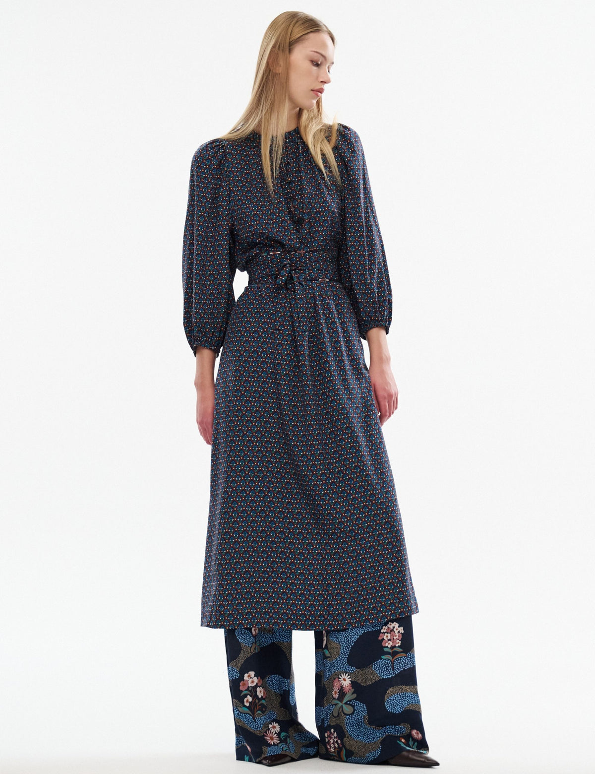 Model wearing a blue patterned belted dress layered over wide-leg floral trousers against a white background.
