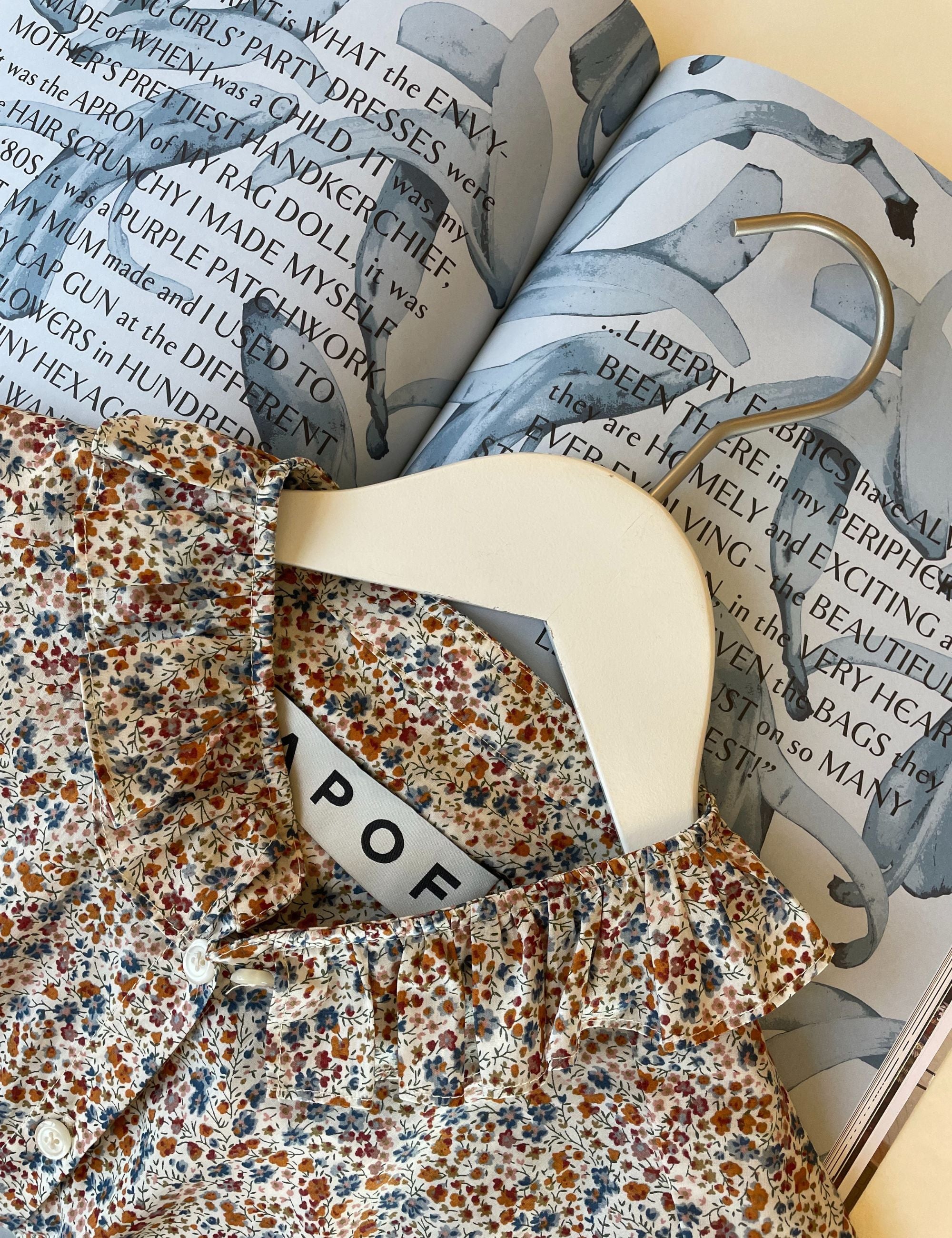 Multicolored floral print shirt with frilled collar on a hanger and blue book about Liberty fabrics