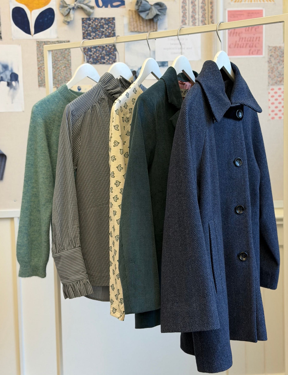 A row of five women's outerwear and knit tops hanging on white hangers from a wooden rack in a boutique. The garments include a sage green sweater, a gray-and-white striped long-sleeve top, a cream floral patterned blouse, a dark green top, and a dark blue textured coat.