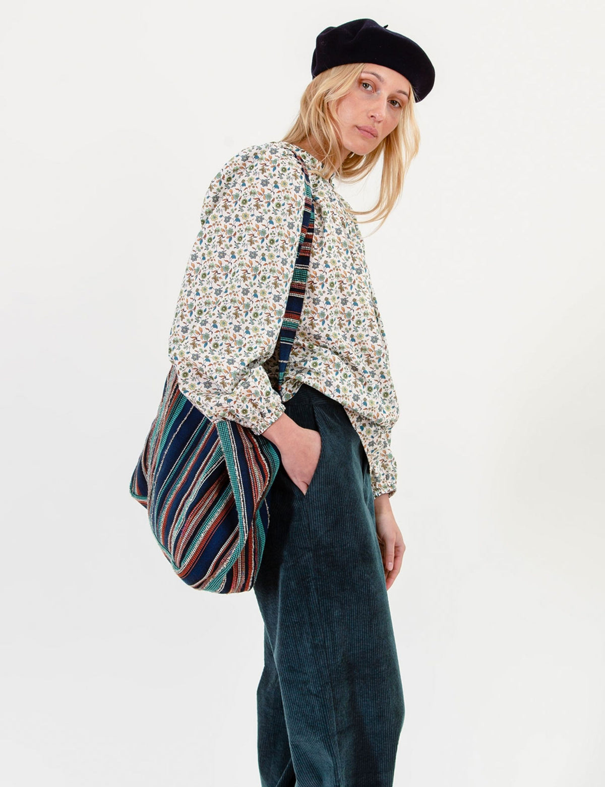 A model wearing a black beret, a long-sleeve floral blouse, and dark green corduroy trousers, carrying a striped cotton shoulder bag against a plain white studio background.