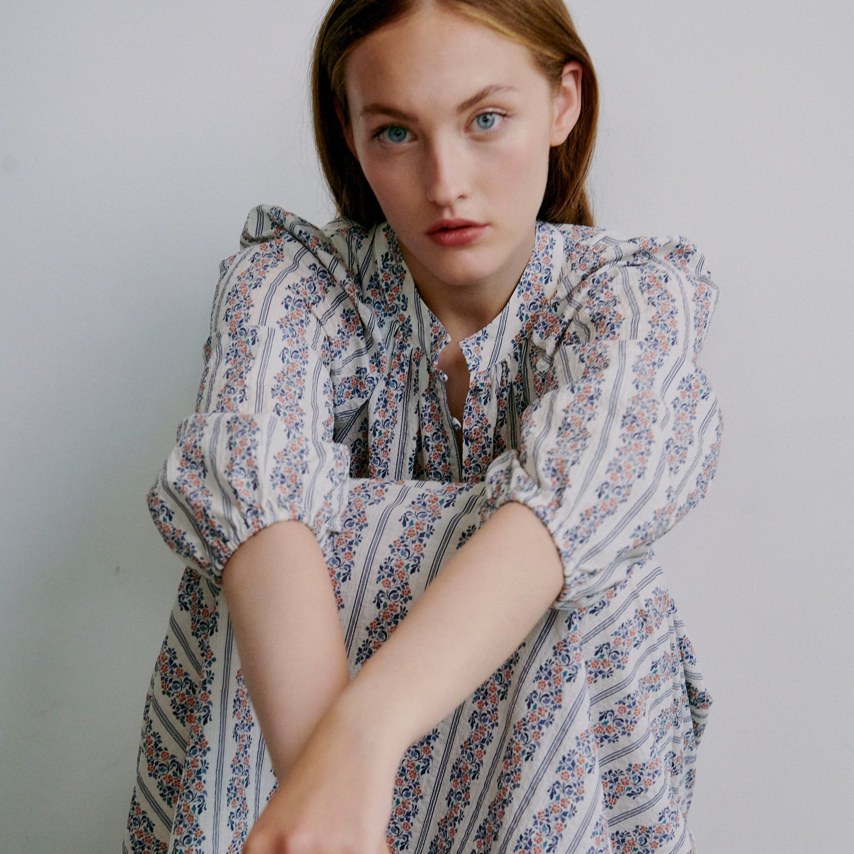 Woman wearing a patterned dress sitting on the floor against a plain background