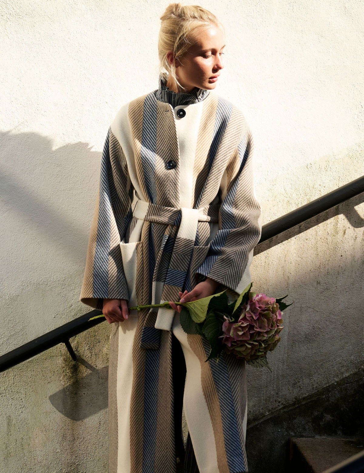 A woman stands on outdoor stairs wearing a long belted coat with a beige, gray, and blue herringbone pattern. She is holding a bouquet of pink flowers against a textured off-white wall.