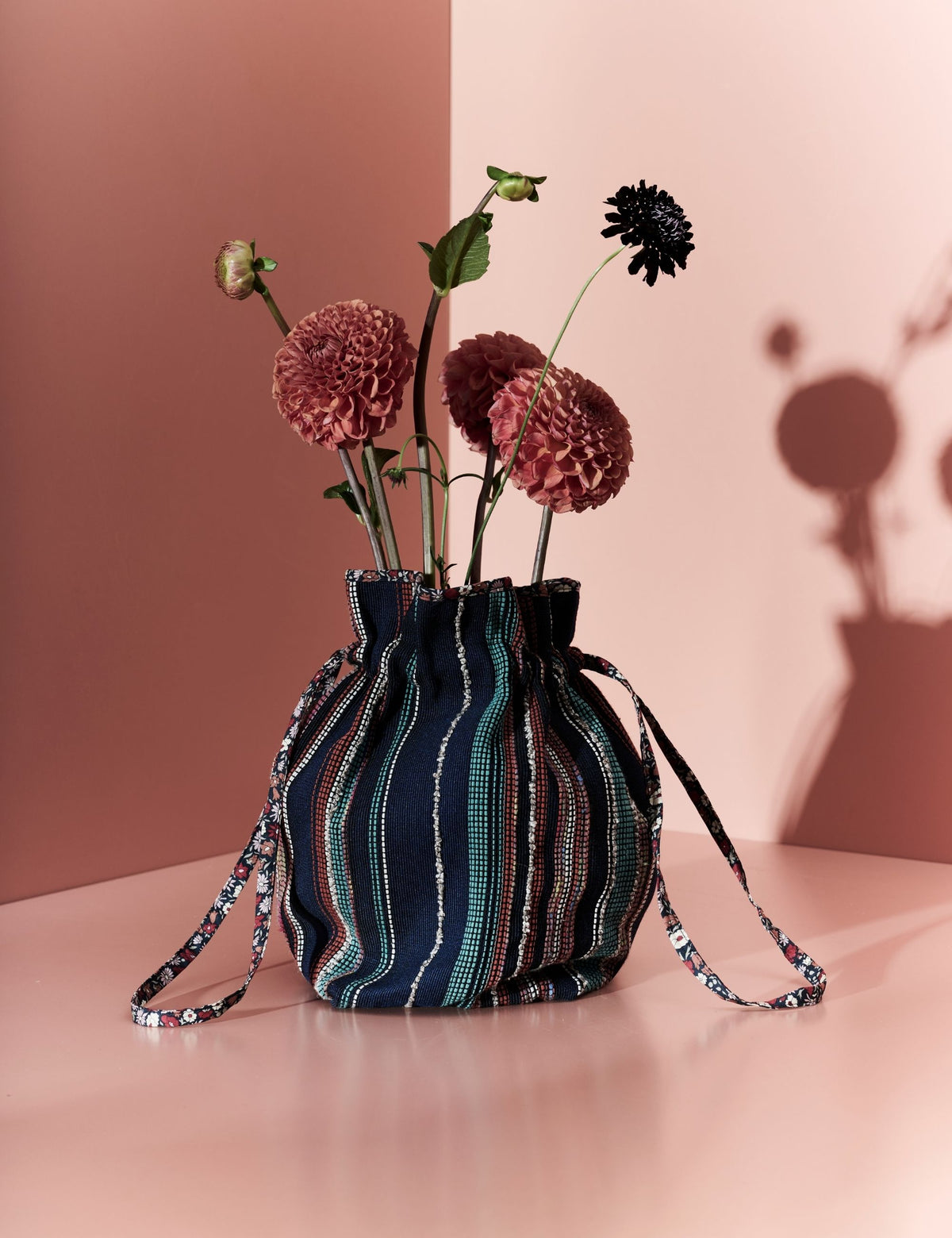 A striped fabric drawstring bag in navy, teal, and cream, styled with dried burgundy flowers on a pink surface against a pink background.