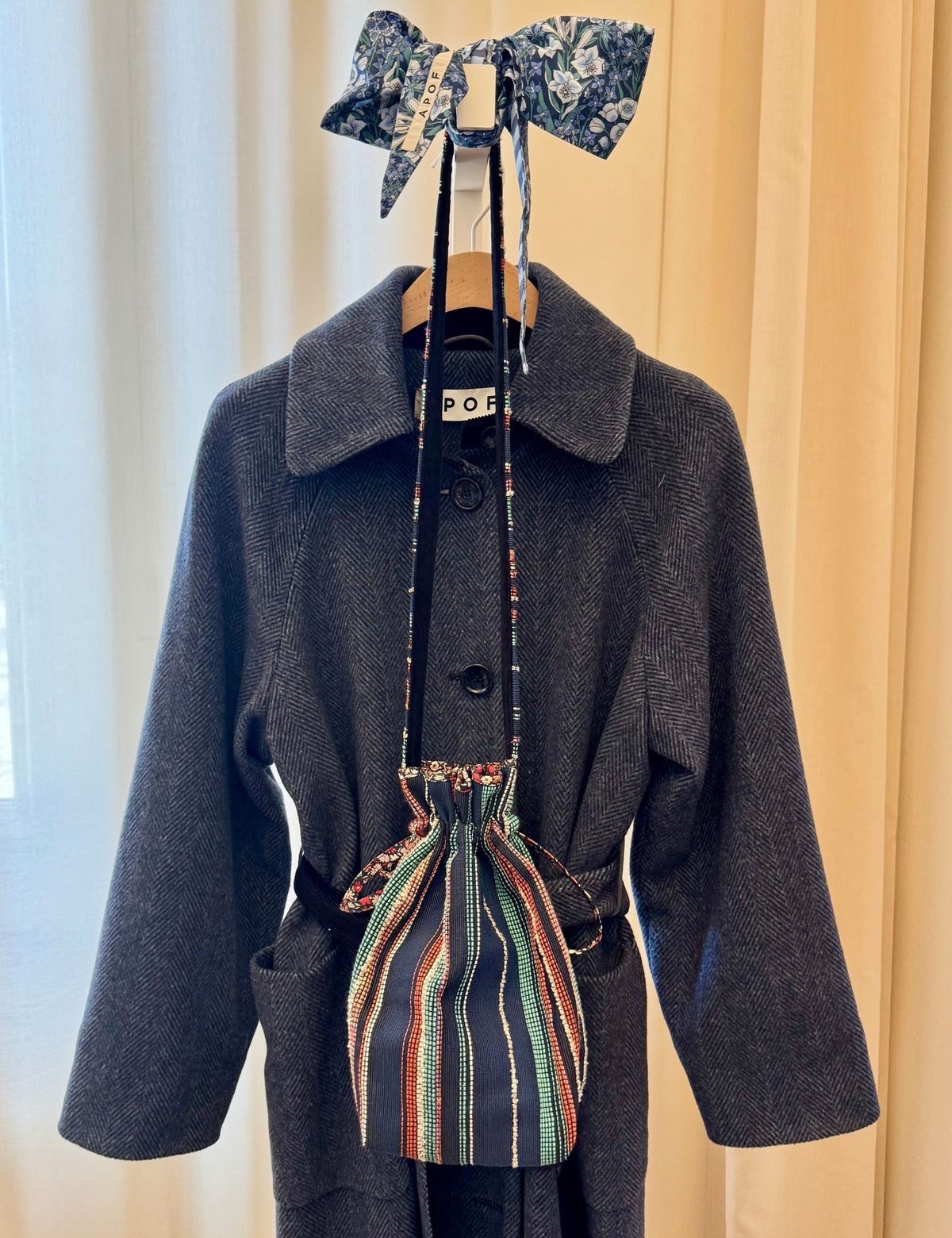 Blue herringbone wool coat styled with a striped drawstring bag and floral bow on a hanger.