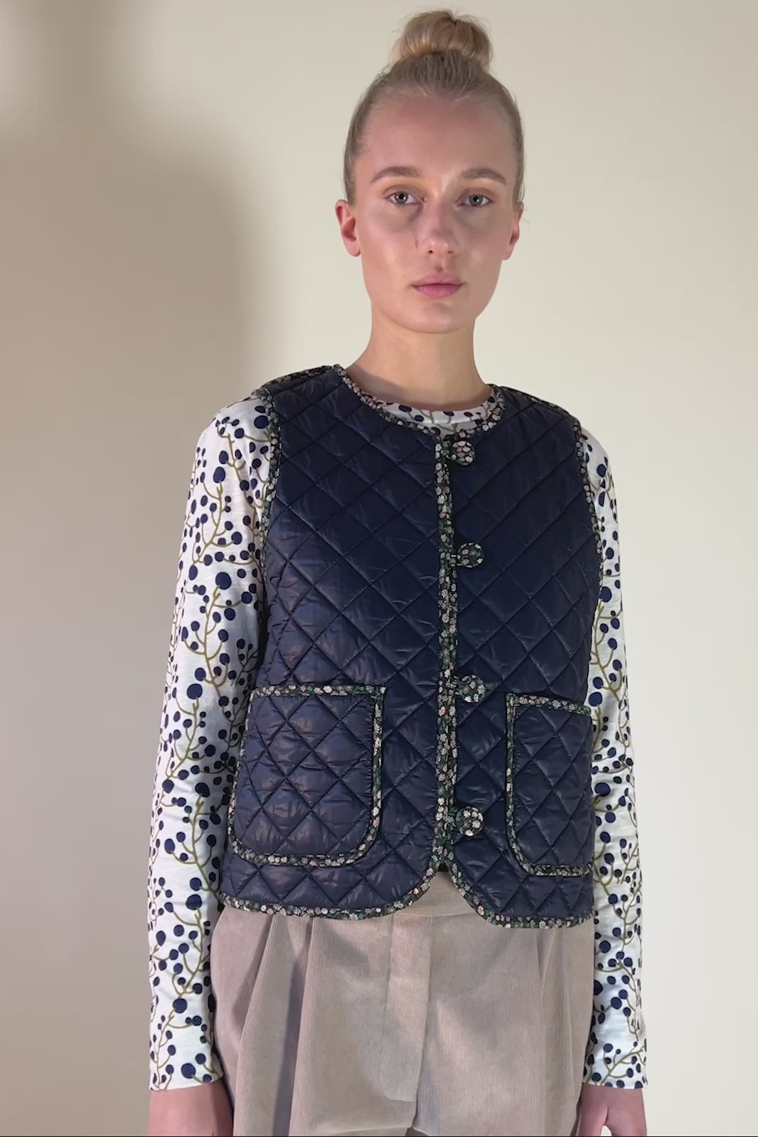 Woman in beige baby corduroy pants, long sleeved t-shirt with blueberry printa an blue quilted vest with buttons and pockets