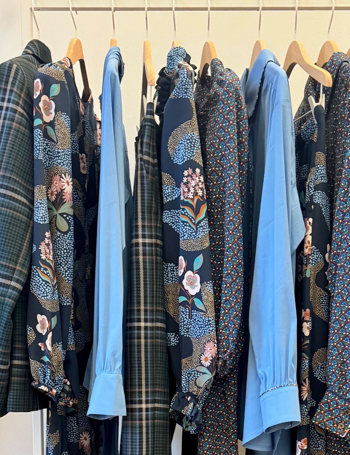 Blue shirts, checked jackets and dark floral prints hanging together on a clothing rack.