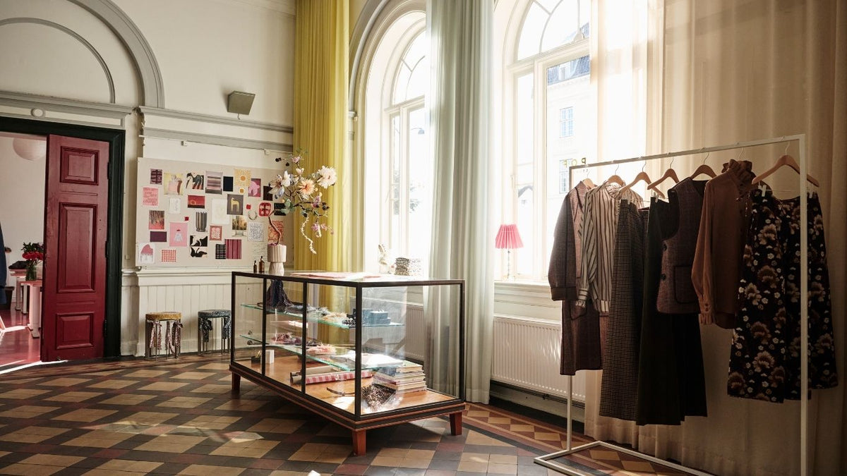 APOF boutique interior with a clothing rack holding several garments, a glass display case, patterned tile flooring, arched windows with yellow and white curtains, and a red door on the left.