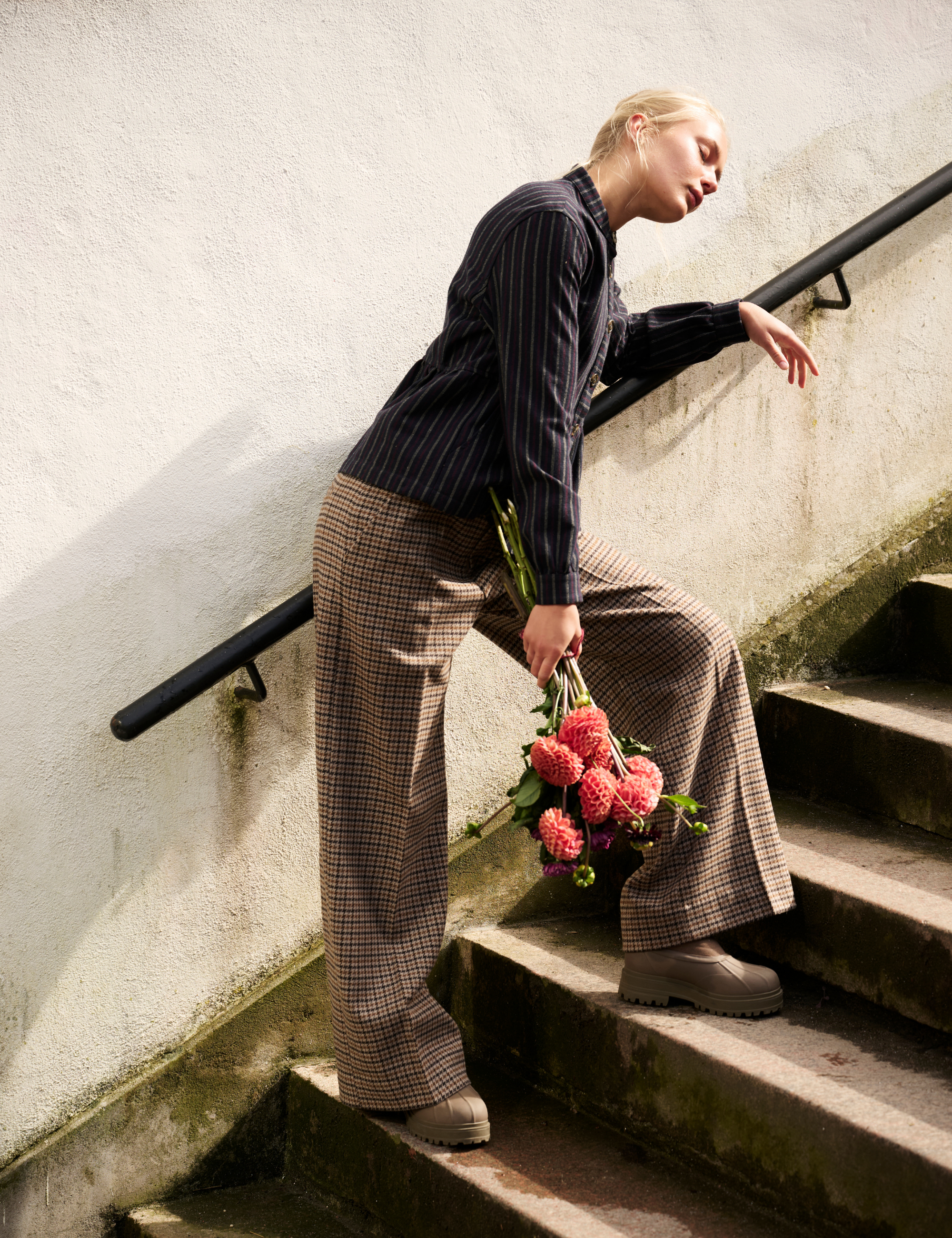 A woman in a dark navy pinstripe shirt and brown plaid wide-leg trousers sits on outdoor concrete stairs against a textured white wall, holding a small bouquet of pink flowers.