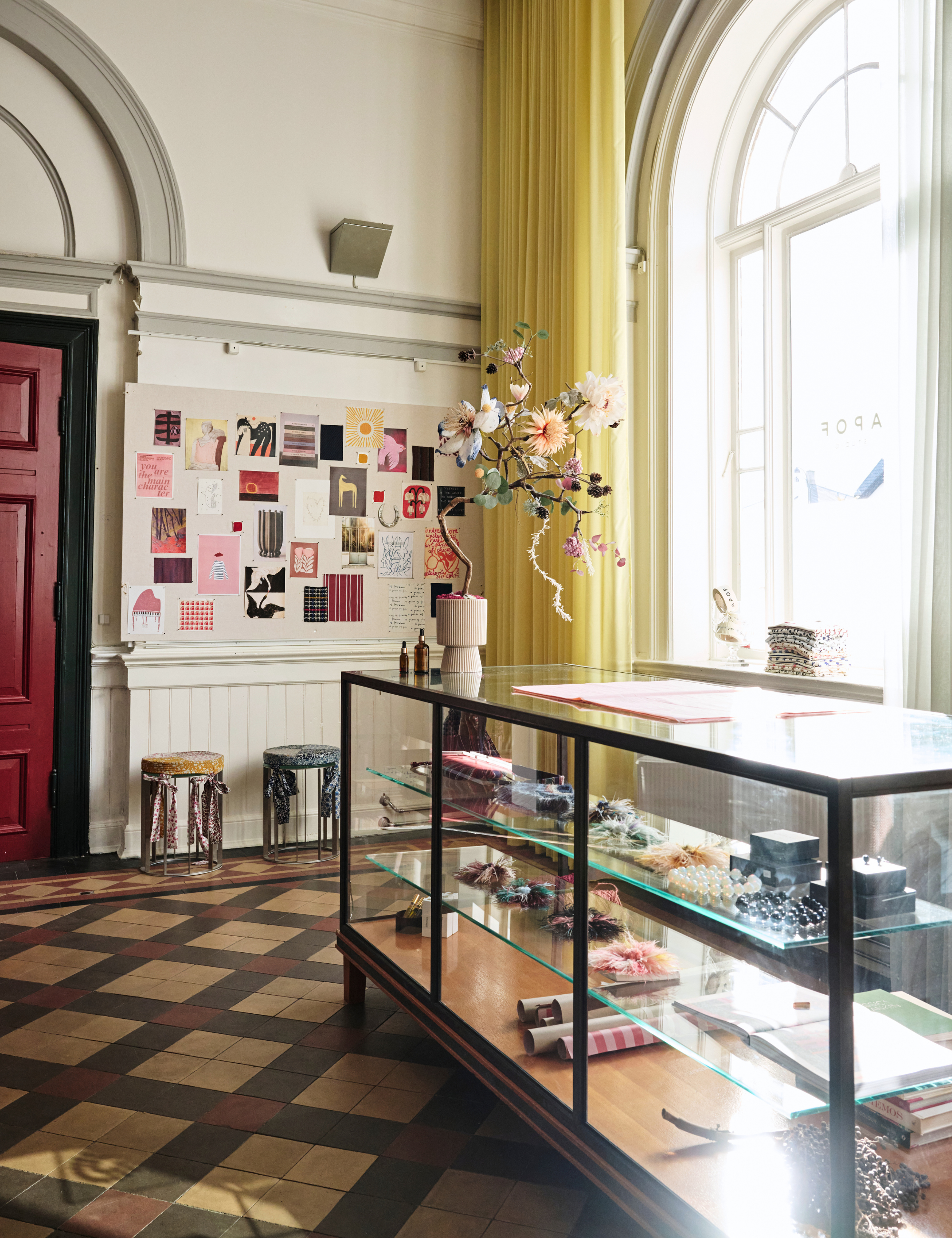 A styled interior scene in a bright room with a glass display case in the foreground, a colorful collage board on the wall, yellow curtains, and a patterned floor.