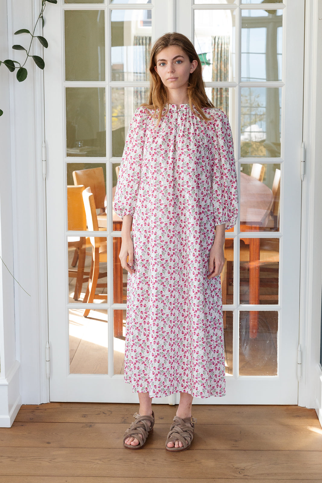 Woman standing at glass door wearing sandals and a long sleeved cotton dress with red small flower pattern and a matching fabric belt